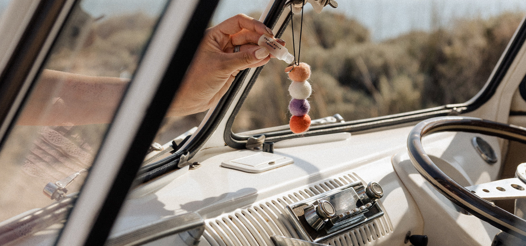 VW Combi van with Smelly Balls reusable air freshener on the rearview mirror with a woman's handing applying essential oil to the felt balls 