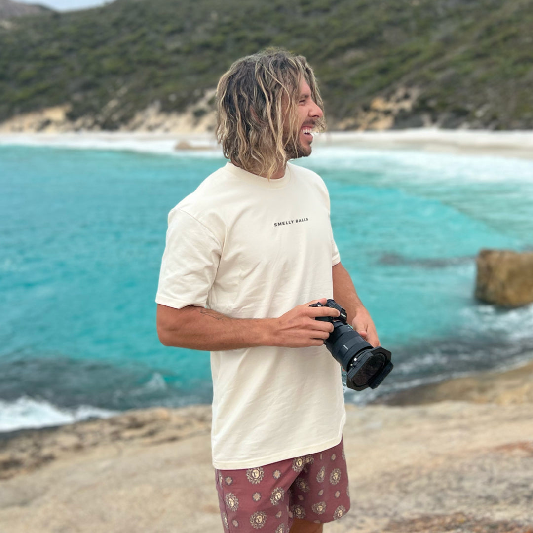 A man standing by the ocean, holding a camera and wearing a "SMELLY BALLS" t-shirt.