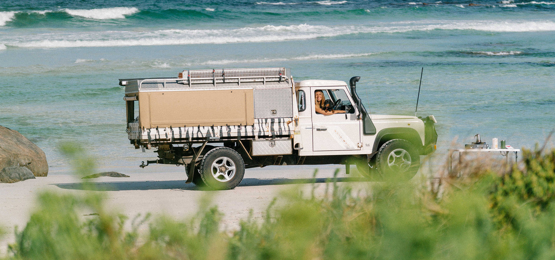 Land Rover Defender with canopy and snorkel, with a woman and a sausage dog smiling out the window with Smelly Balls reusable air freshener hanging in the rearview mirror on the beach with a camp table with a camp cooker and espresso maker 