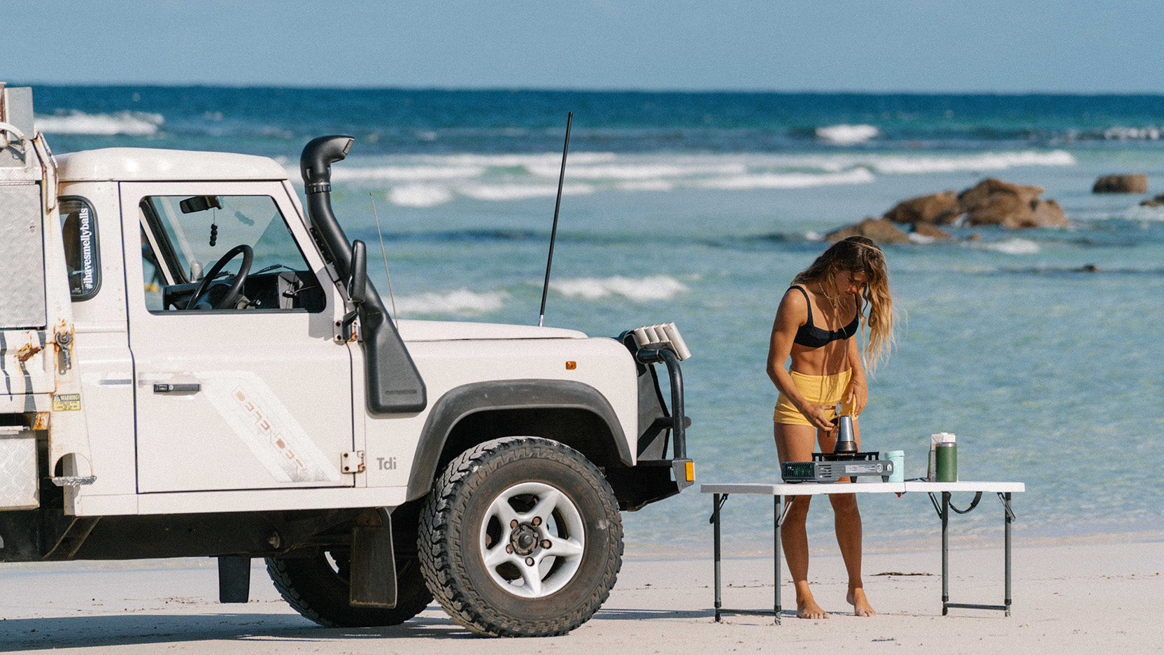 A landcruiser defender with a canopy and snorkel and Smelly Balls reusable air freshener on the rearview mirror on the beach with a woman in a swimsuit using a espresso maker to make a cup of coffee