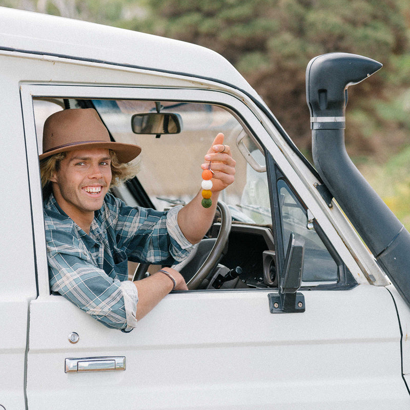 Man sitting in a car holding a colorful string of balls, smiling and wearing a hat.