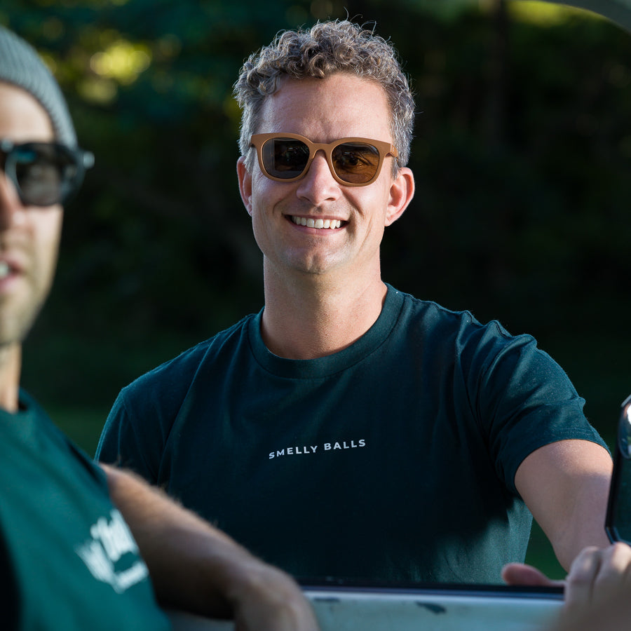 A man wearing sunglasses and a "Smelly Balls" t-shirt smiles.